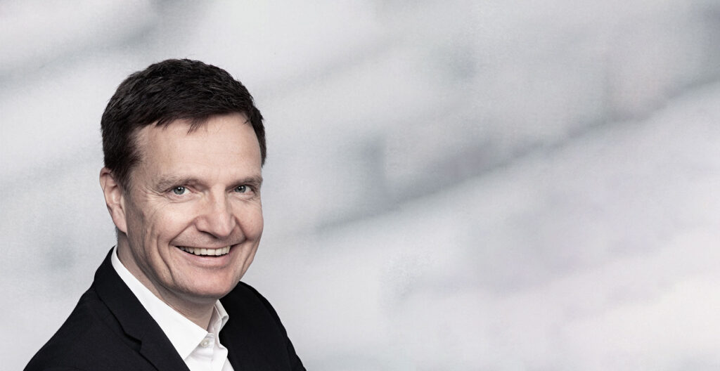 Moderator Dirk Böge im schwarzen Sakko mit weißem Hemd lächelt.