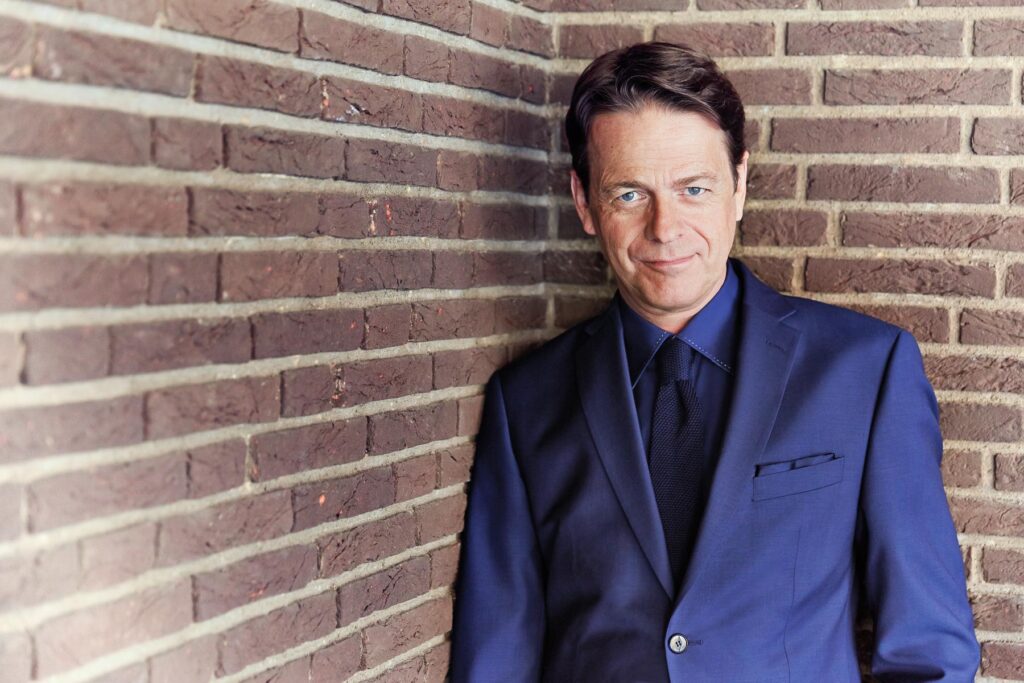 ZDF Moderator Rudi Cerne lehnt im blauen Jacket mit blauem Hemd vor einer Backsteinwand.