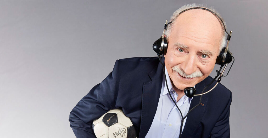 Moderator Werner Hansch mit antikem Kommentatoren-Headset im blauen Jacket und hellblauem Hemd mit Fußball unter dem Arm.