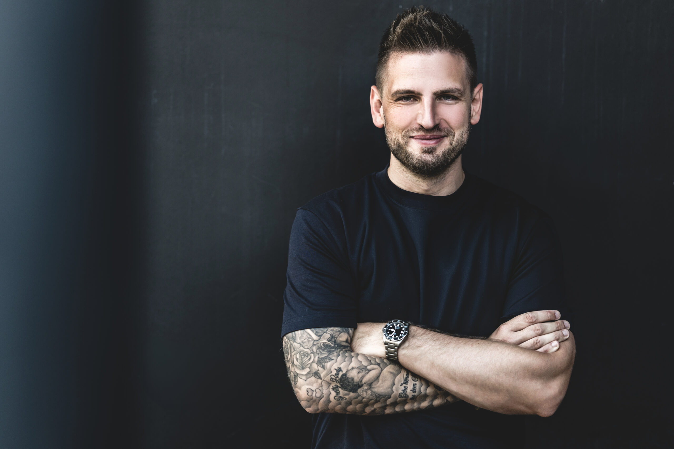 Moderator Uli Hebel im schwarzem T-Shirt mit verschränkten Armen vor einer dunklen Wand.