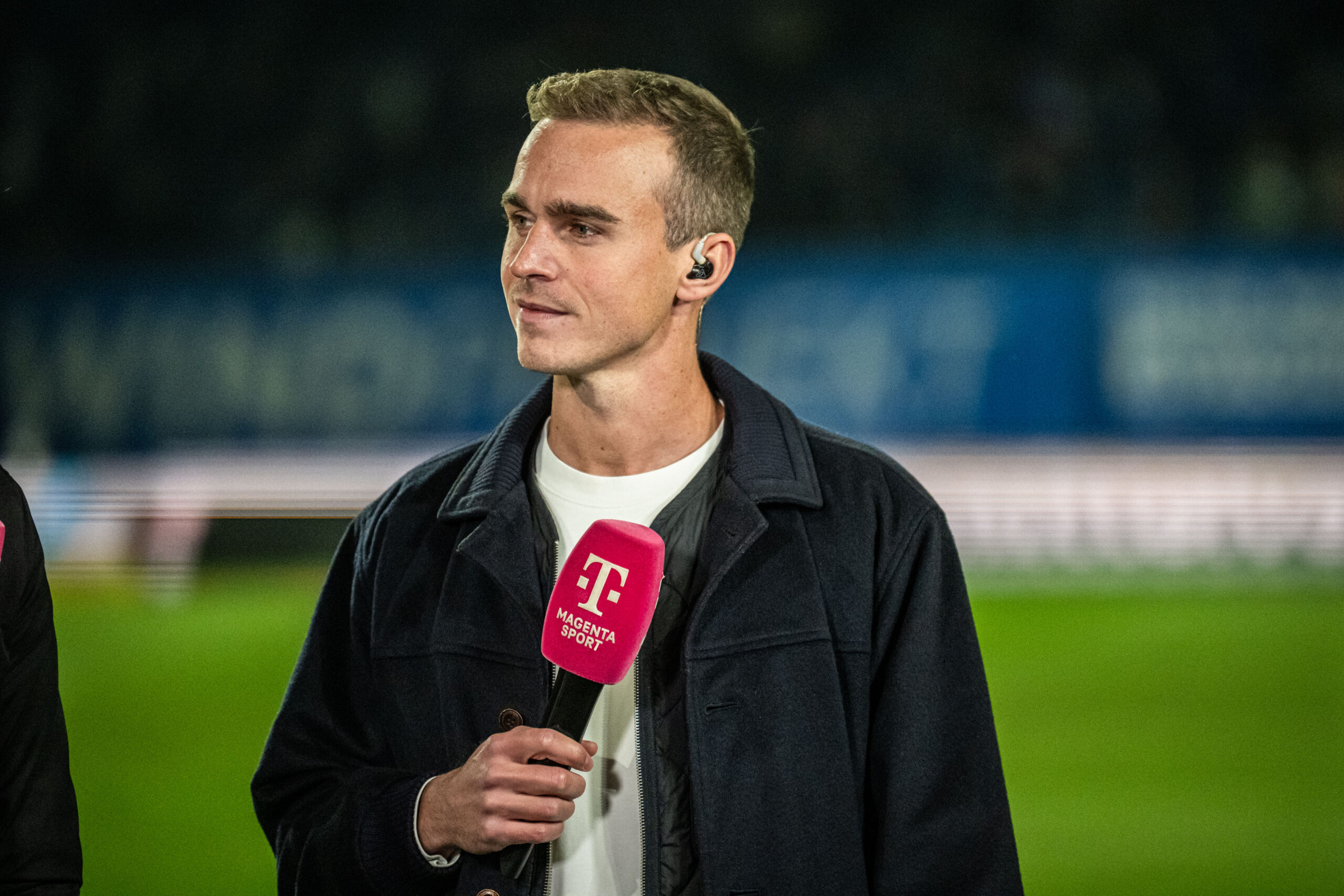 Moderator Hannes Jakobi steht mit blauer Jacke, weißem T-Shirt und Magenta Mikro in der Hand auf dem Rasen eines Fußballstadions.